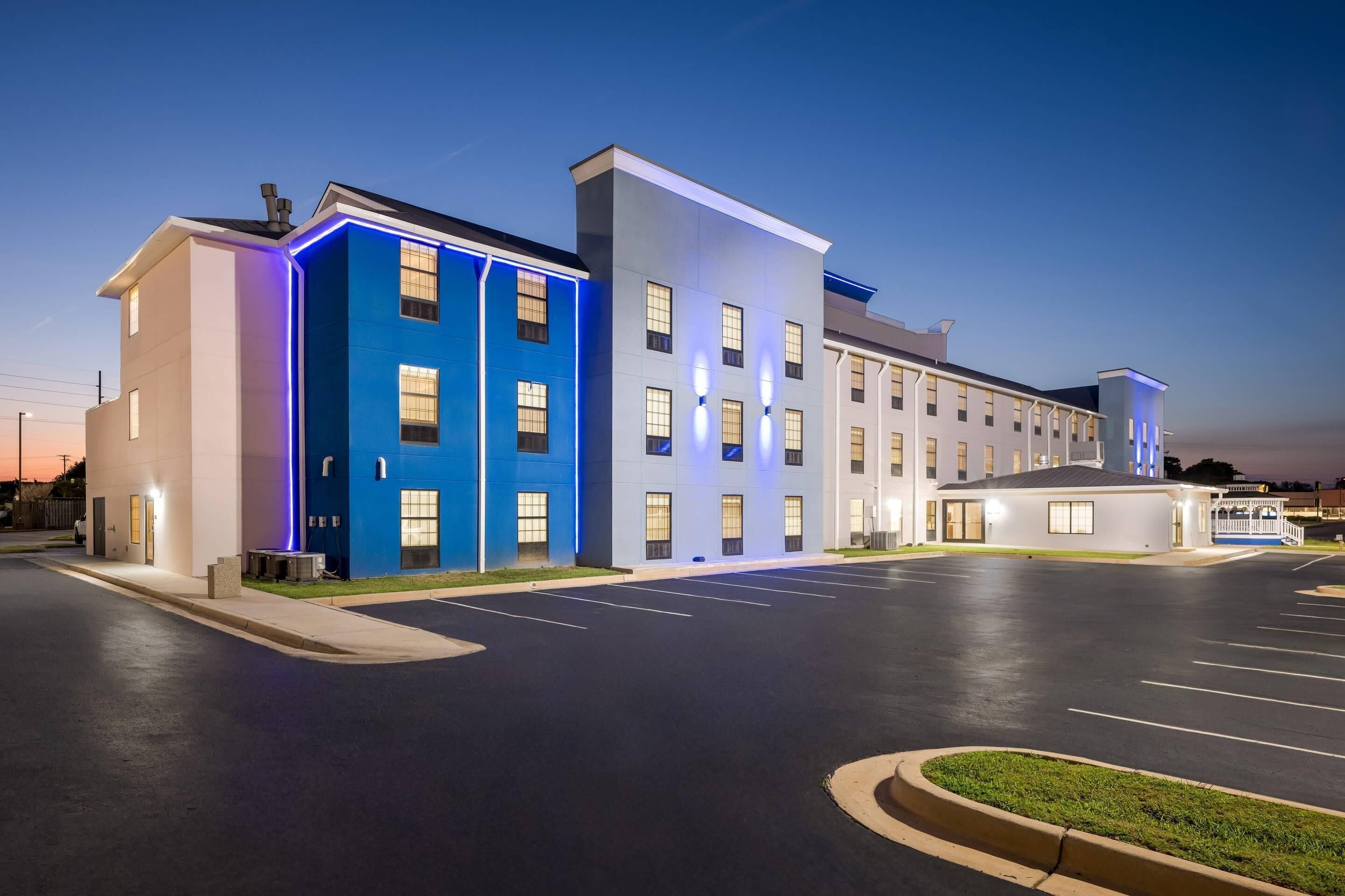 Hotel moderno com entrada iluminada em azul, amplo estacionamento e fachada bem iluminada.