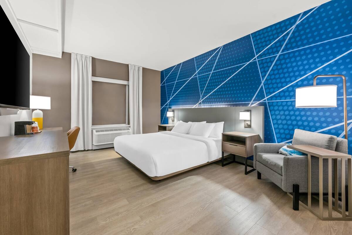 A modern hotel room with a large bed, a desk, a chair, and a TV.