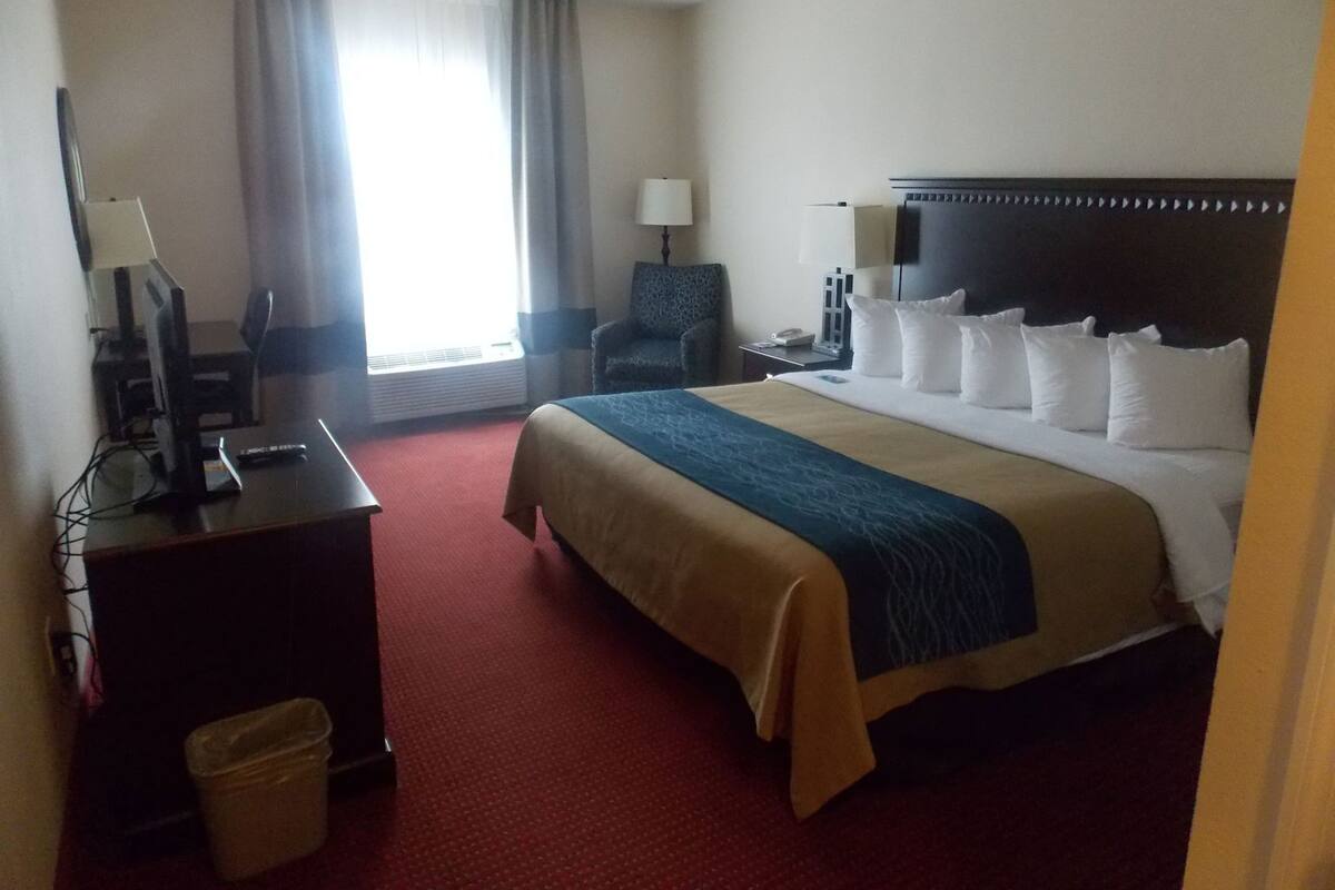 A hotel room with a bed, a desk with a computer, a chair, a lamp, and a window with curtains.