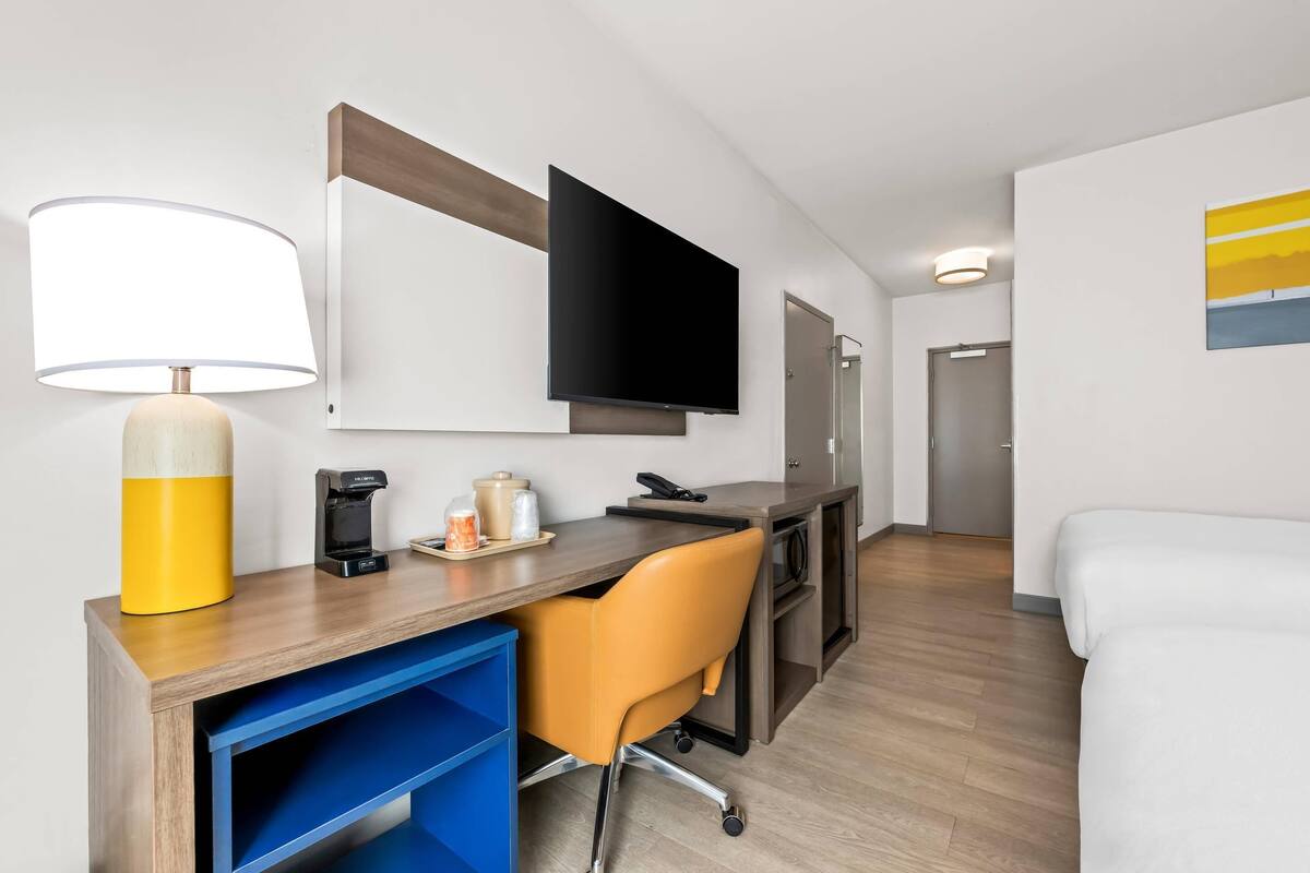 A modern hotel room with a wooden desk, a yellow lamp, a flat-screen TV, a coffee maker, and a white bed.