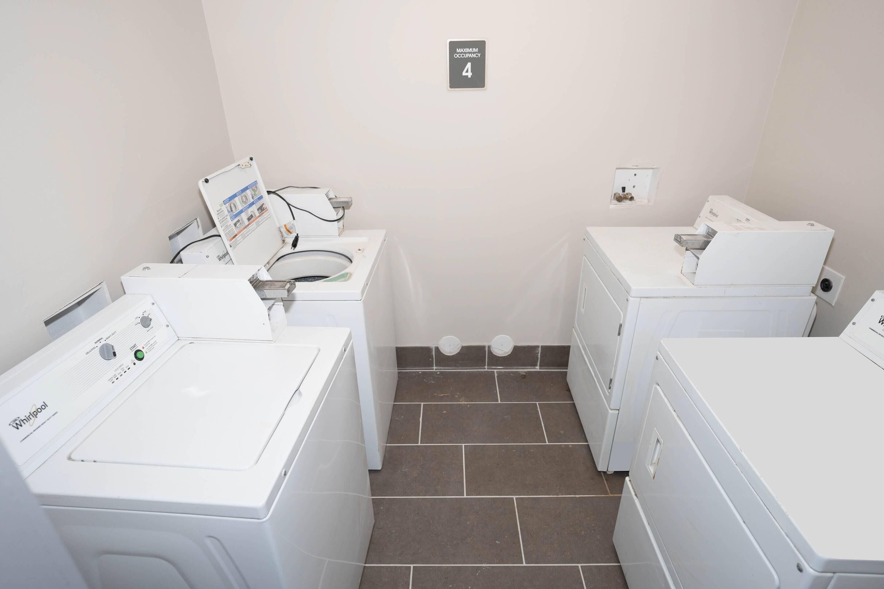 A laundry room with four Whirlpool washers and dryers, a sign indicating a maximum occupancy of 4, and a tiled floor.