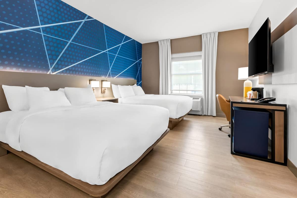 A hotel room with two beds, a desk with a computer, a TV, and a large window with curtains.