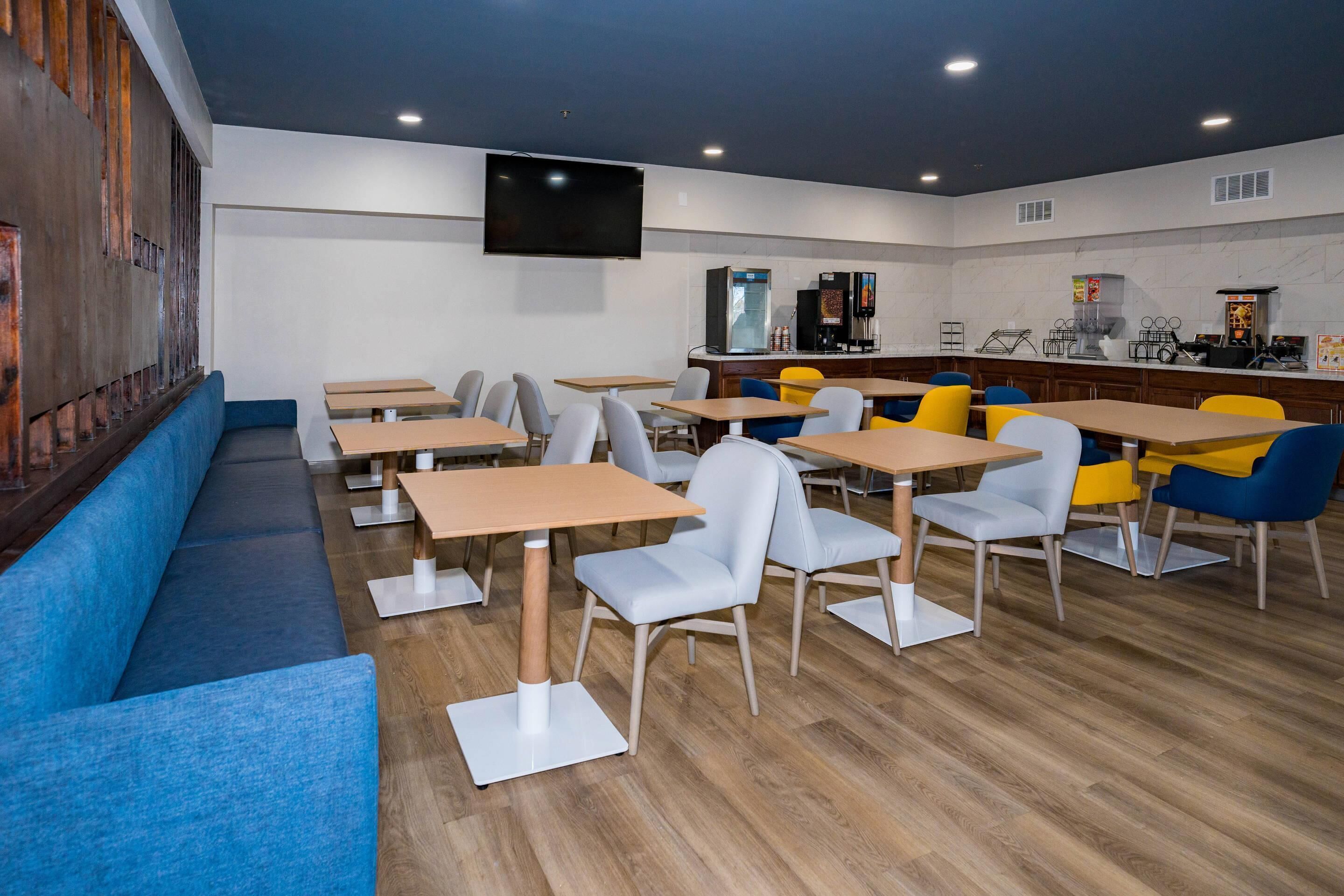 A modern restaurant with wooden tables and chairs, a flat-screen TV, and a bar area with a coffee machine.
