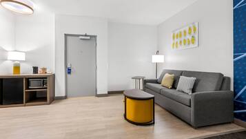 Una habitación de hotel moderna con un sofá gris, una mesita redonda amarilla, un microondas, una lámpara y una puerta.