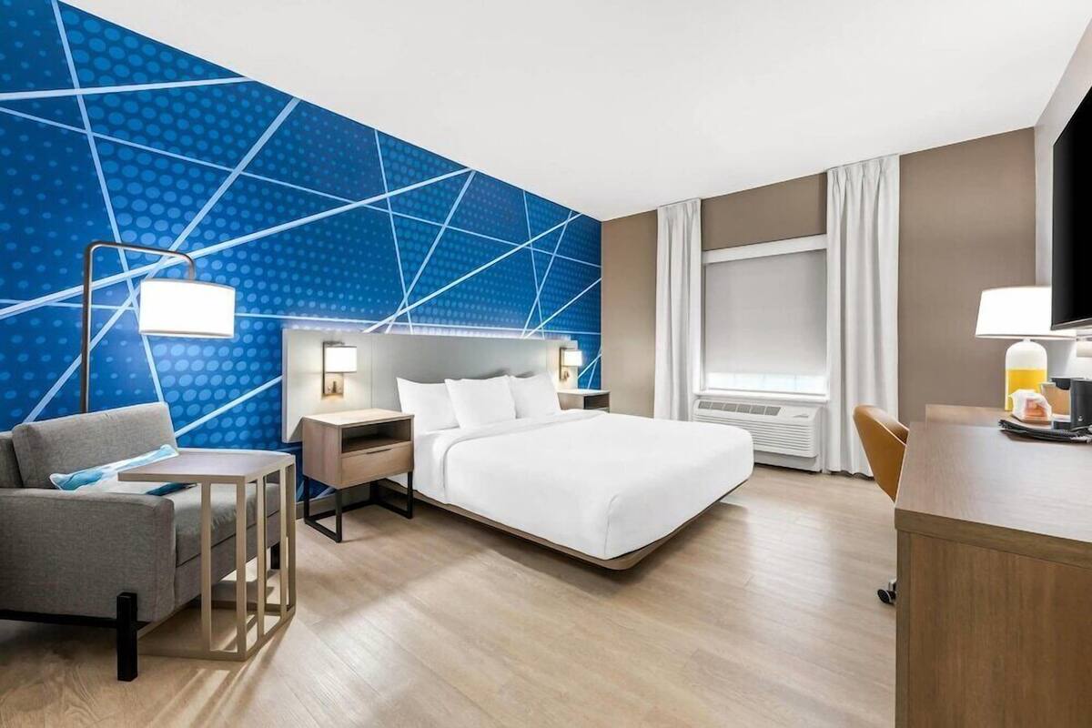 A hotel room with a bed, a desk, a chair, a nightstand, and a wall with a geometric pattern.