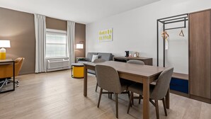 A modern living room with a dining table, sofa, and a small side table. There is a window with curtains, a lamp, and a coat rack with clothes. - Comfort Inn & Suites River City Decatur (Decatur)