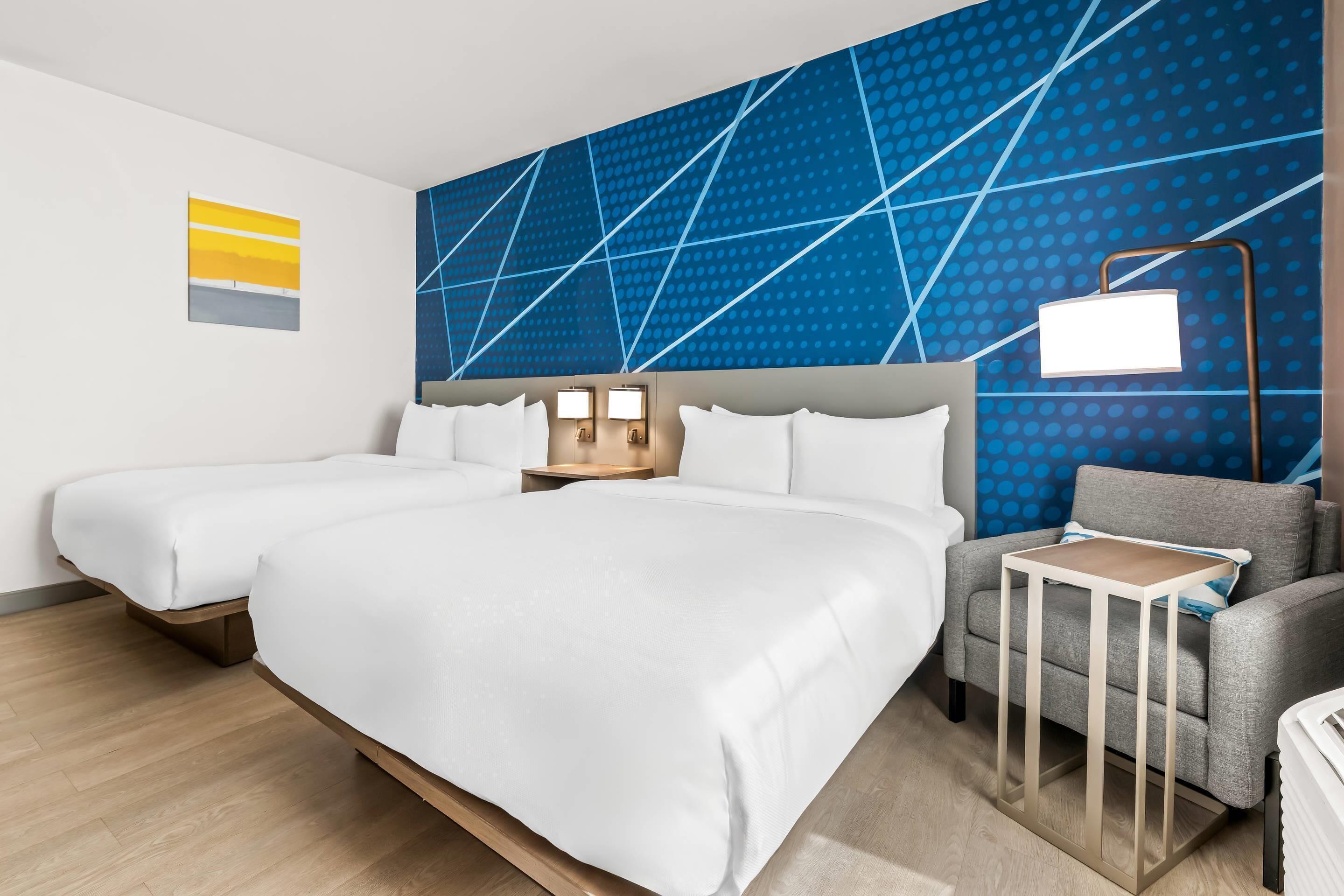 A hotel room with two beds, a grey armchair, a small table, and a wall with a geometric pattern.