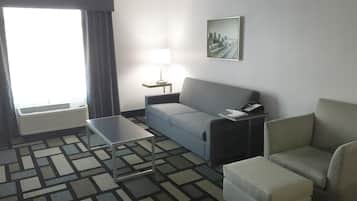 Habitación de hotel con sofá, una mesita, una silla, lámpara, teléfono y un cuadro de un paisaje urbano en la pared.