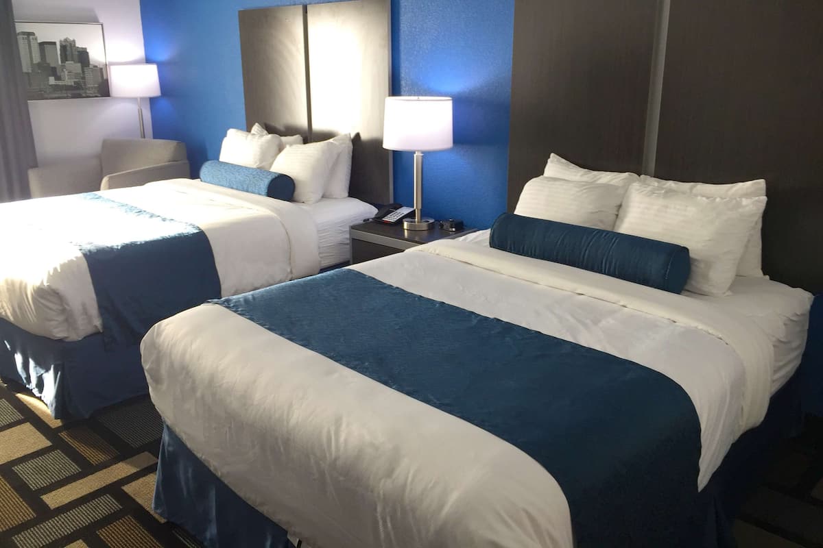 Una habitación de hotel con dos camas, cada una con ropa de cama blanca y azul, una mesita de noche con lámpara y un espejo que refleja un paisaje urbano.