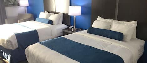 Una habitación de hotel con dos camas, cada una con ropa de cama blanca y azul, una mesita de noche con lámpara y un espejo que refleja un paisaje urbano.