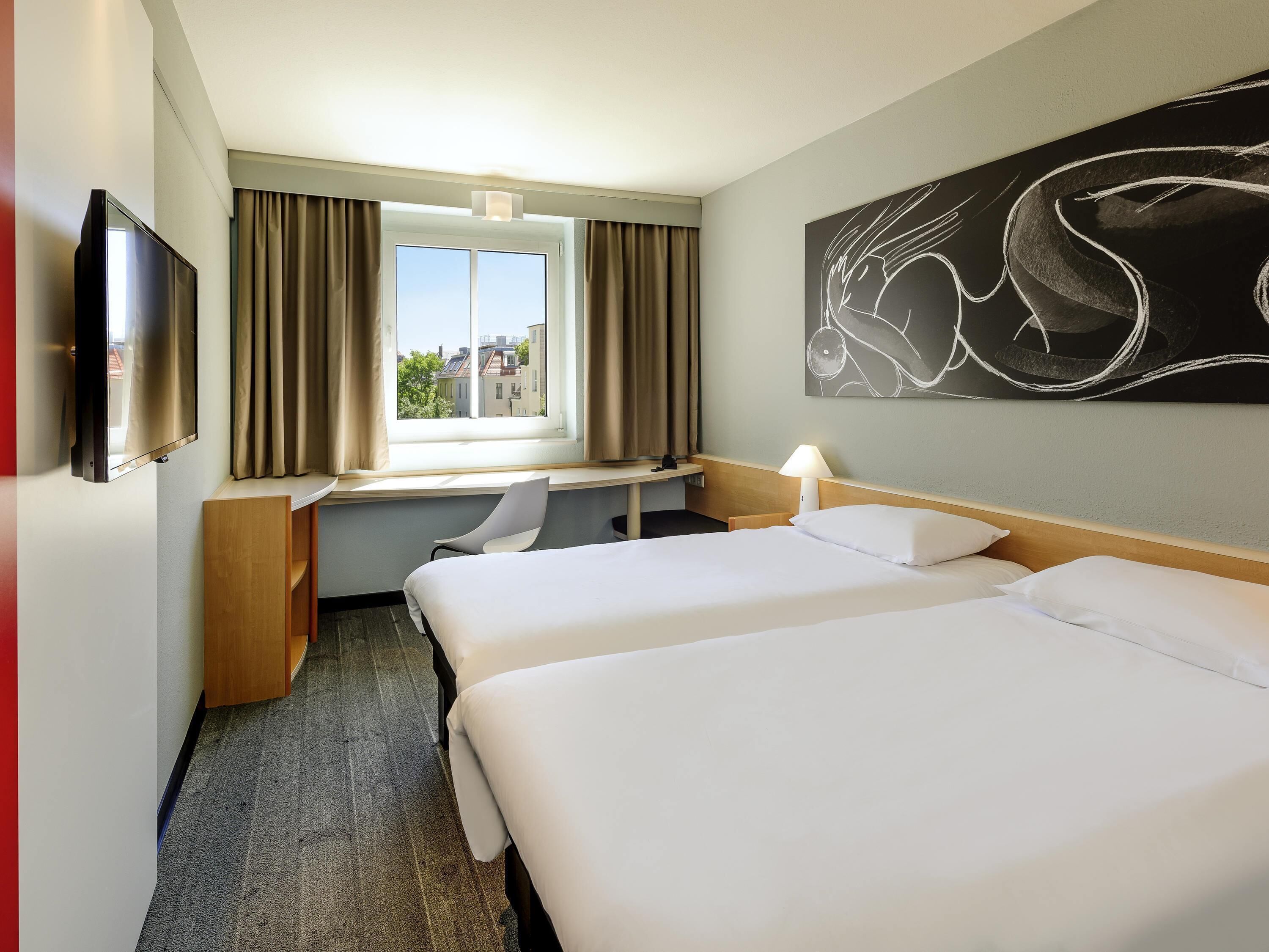 Habitación de hotel con dos camas, un escritorio, una silla, un televisor y un ventanal con cortinas.