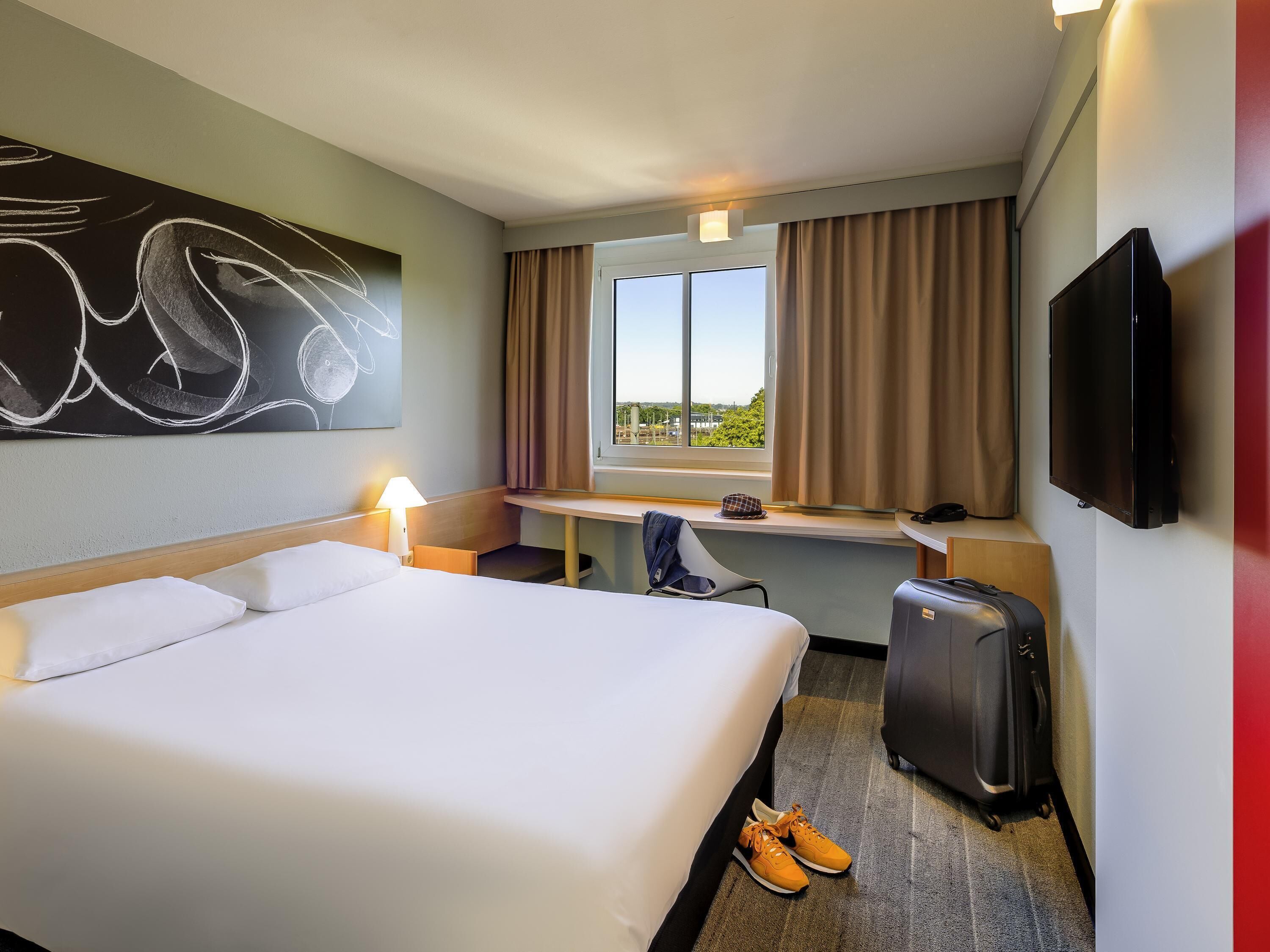 A hotel room with a bed, desk, chair, TV, and a suitcase.