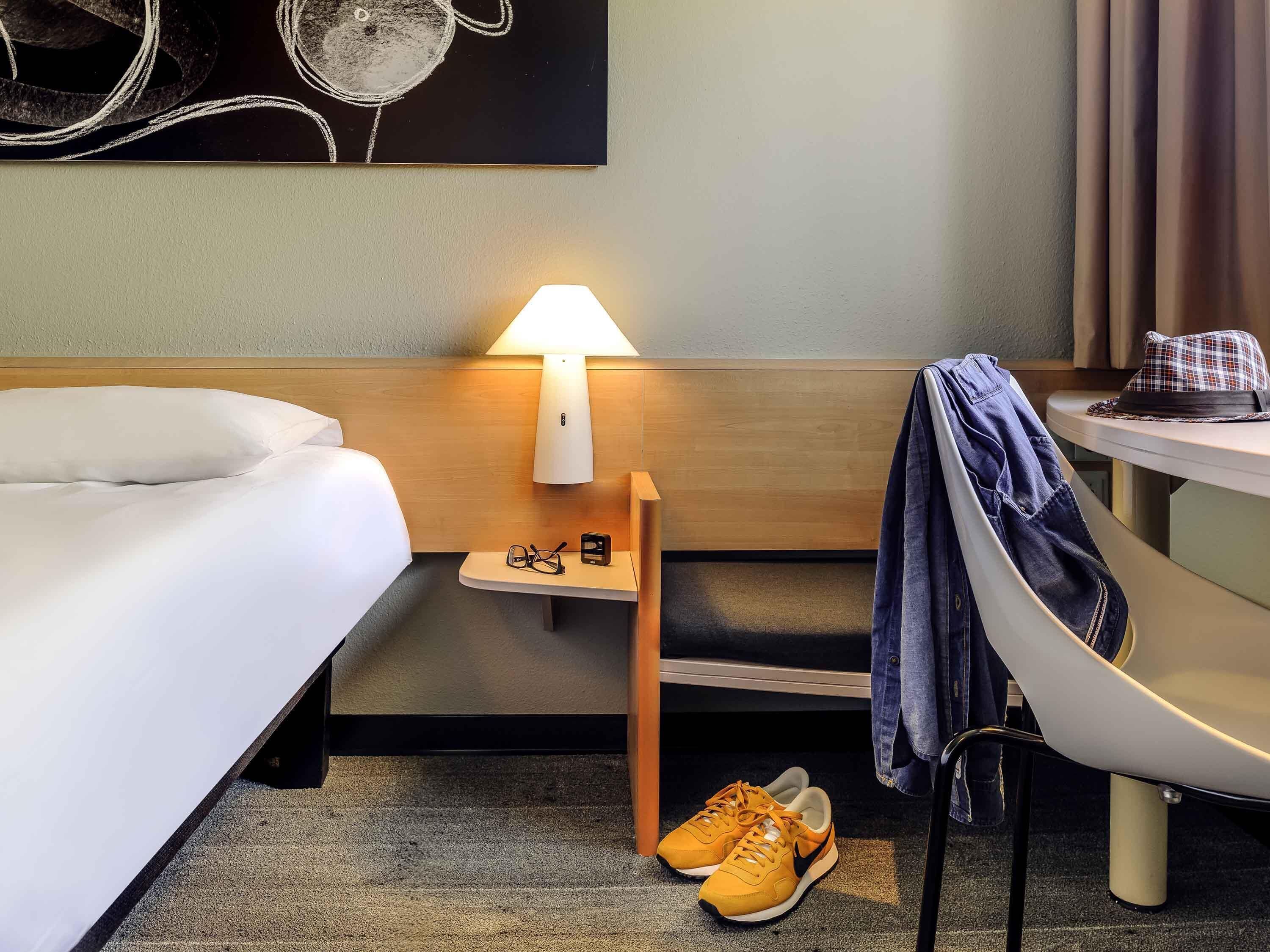 Una habitación de hotel con cama, mesita de noche con lámpara, un par de zapatos y una silla.