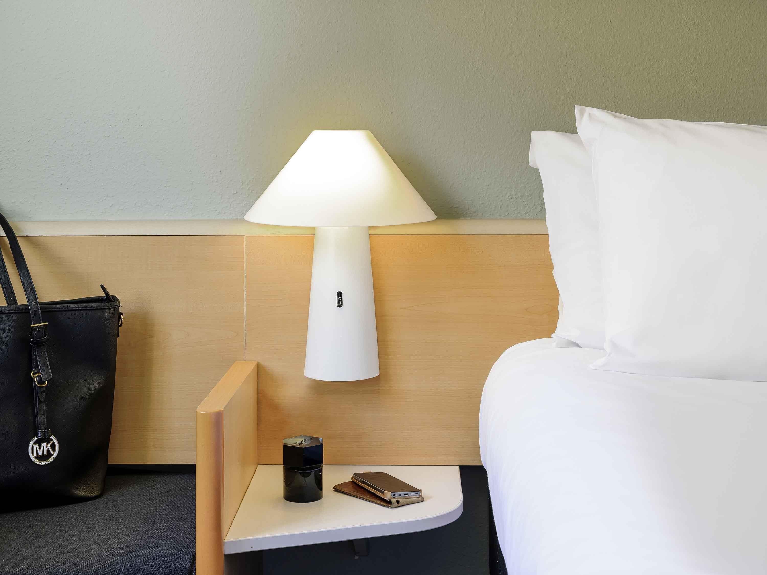 Una habitación de hotel con una cama, una mesita de noche con una lámpara, un bolso negro y un teléfono móvil.