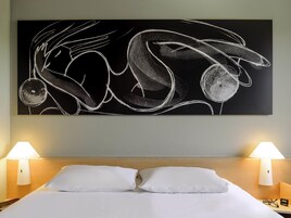 Una habitación de hotel con un cuadro abstracto grande, dos lámparas de noche y una cama bien hecha con ropa blanca.