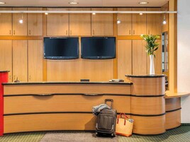 Un mostrador de recepción con paneles de madera, dos televisores de pantalla plana, una planta en maceta, un bolso y una maleta.
