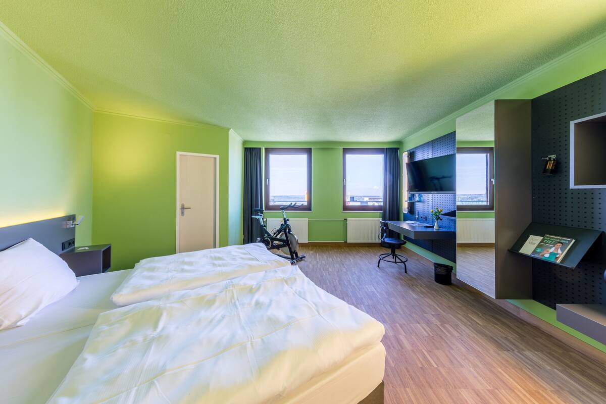Quarto de hotel com cama, escrivaninha, bicicleta e vista para a cidade.
