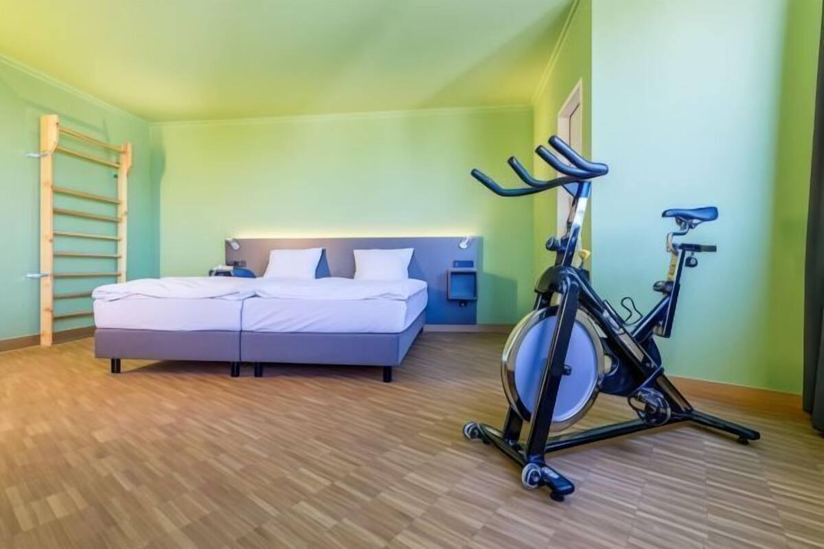 Quarto com cama, bicicleta ergométrica na parede e escada de madeira.