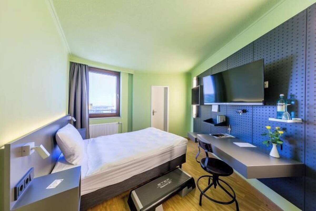 Quarto de hotel com cama, escrivaninha, cadeira, televisão e janela com cortinas.