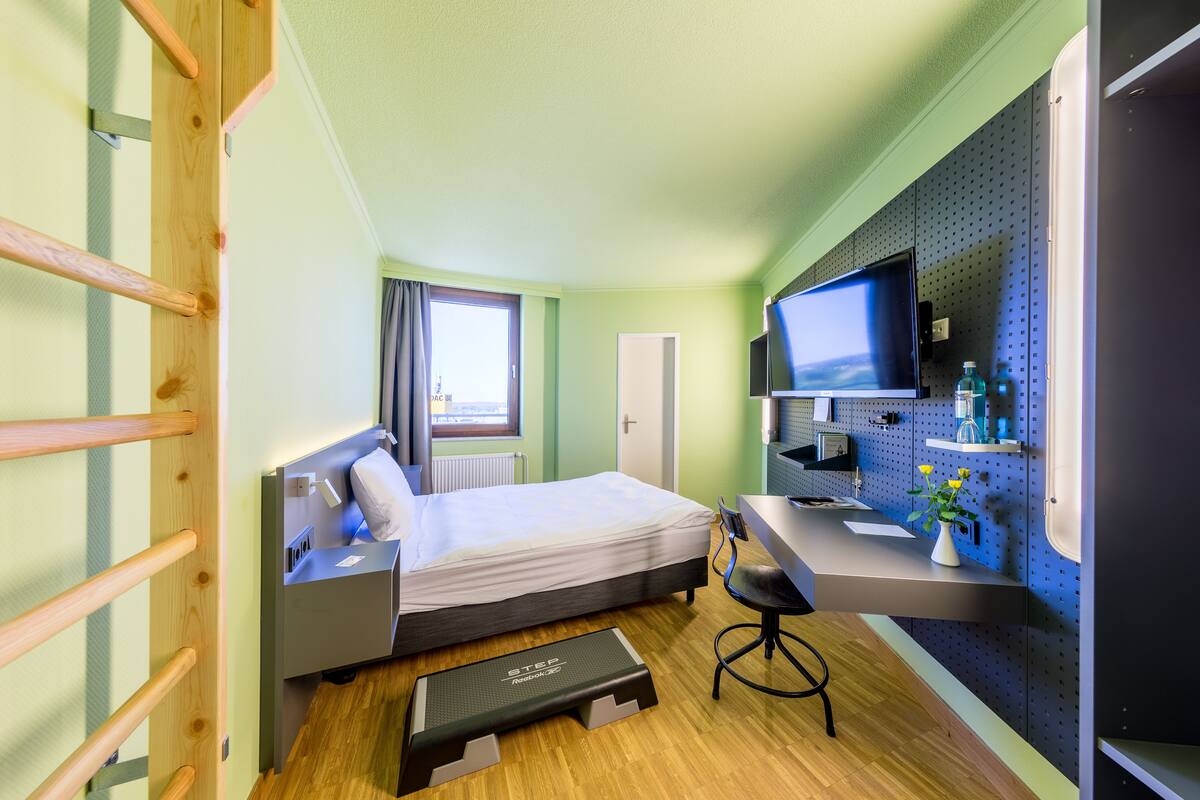 Quarto de hotel compacto com cama, escrivaninha com computador, televisão e uma pequena janela.
