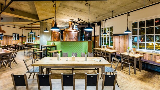 Ein geräumiges Restaurant mit Holztischen und -stühlen, hängenden Pendelleuchten und einem mit grünen Fliesen ausgekleideten Barbereich.