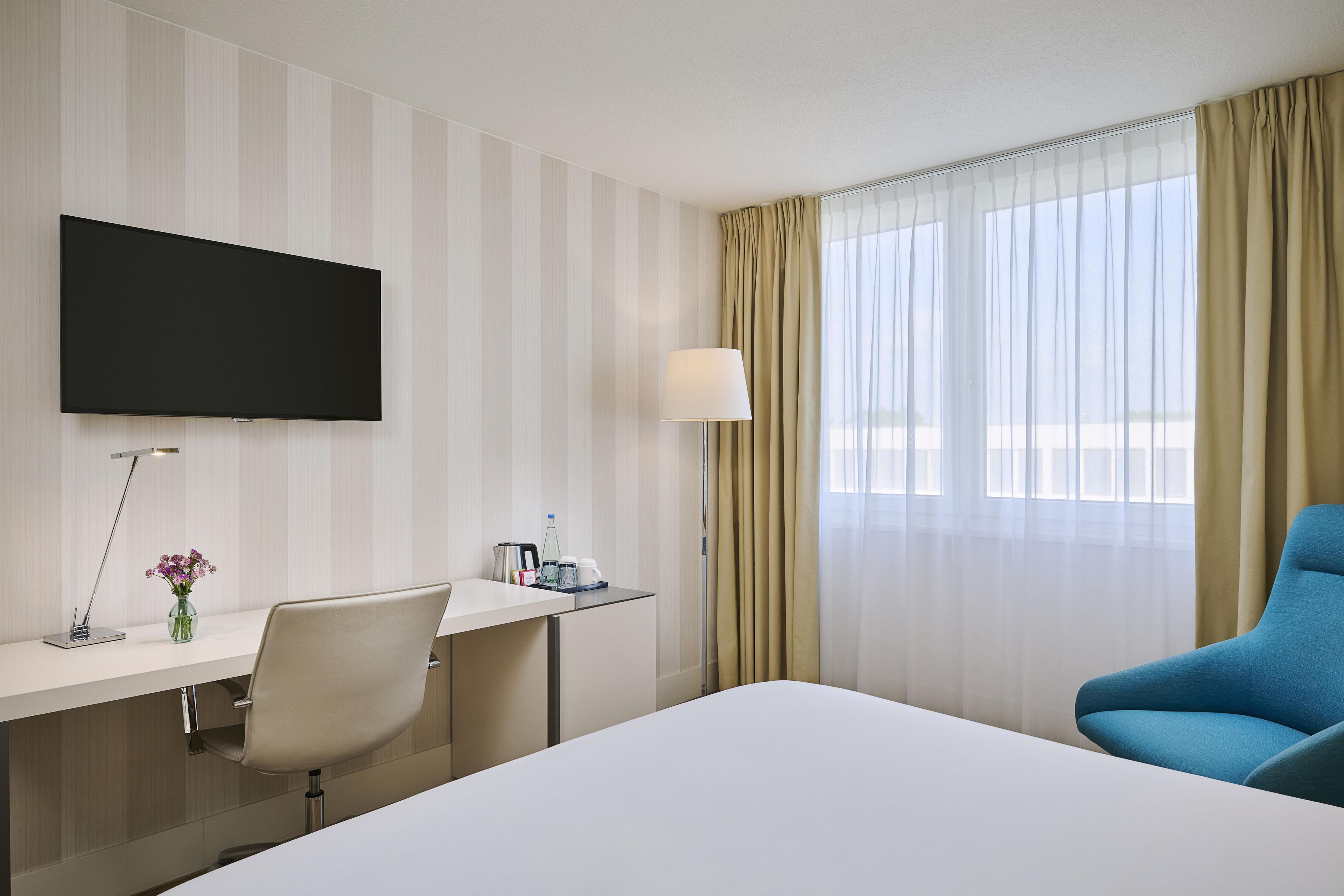 Une chambre d’hôtel avec un lit, un bureau, une chaise, une télévision et une fenêtre avec des rideaux.