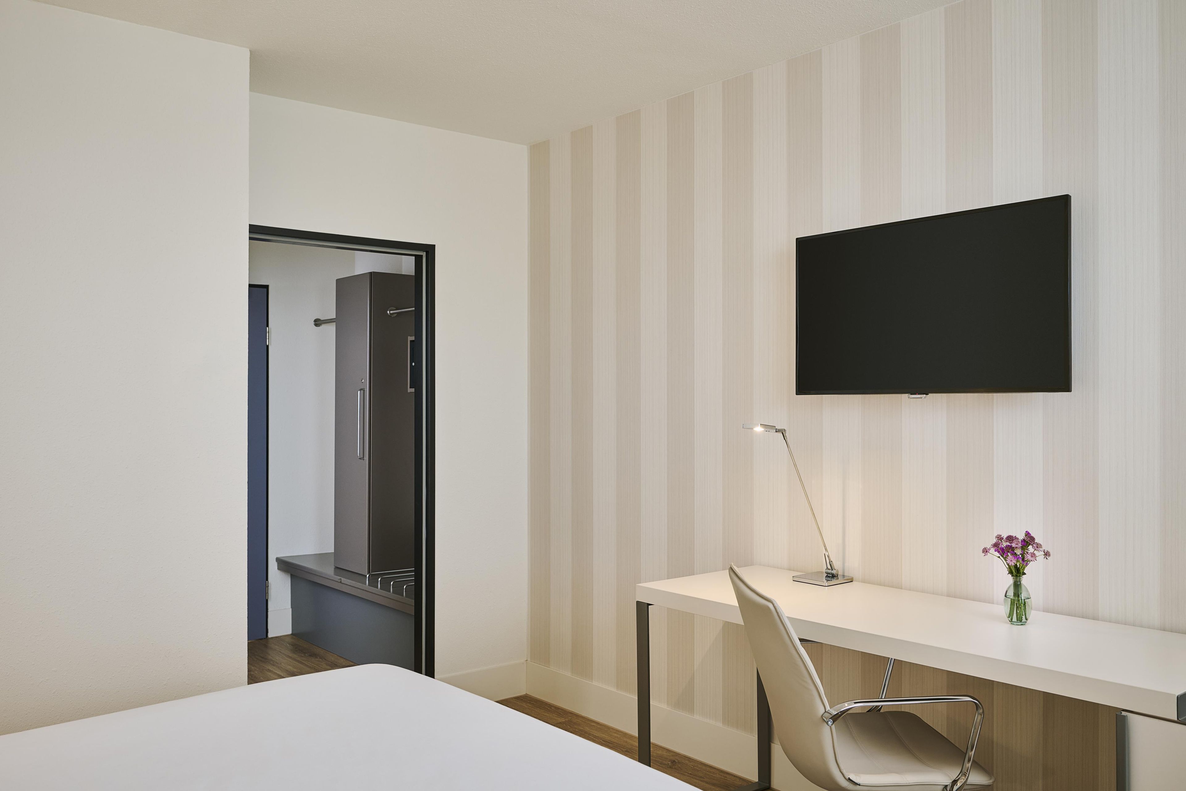 Ett hotellrum med en säng, ett skrivbord med en stol, en platt-TV och en spegel.