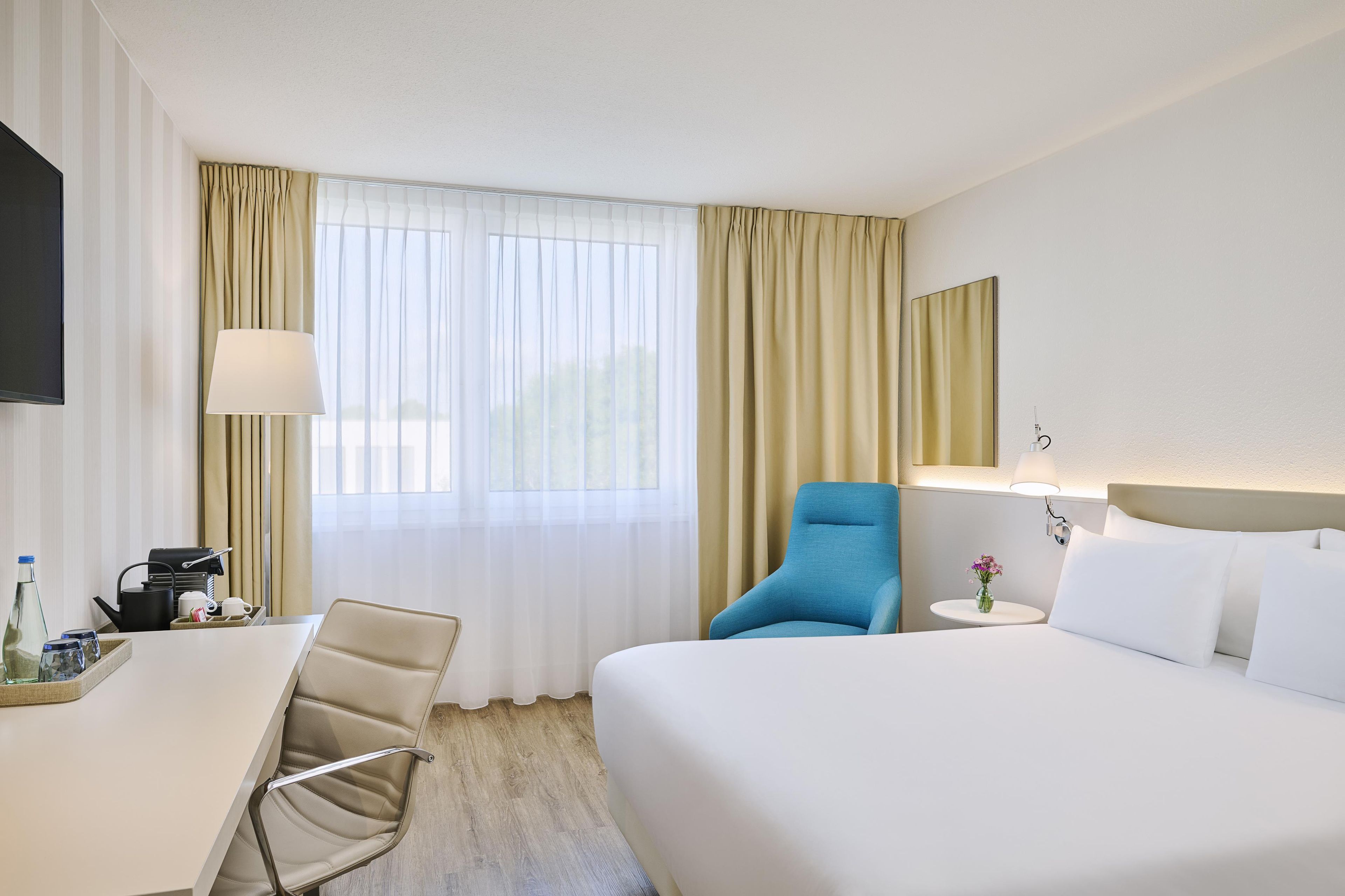A hotel room with a bed, a desk, a chair, a small table with a vase of flowers, and a television.