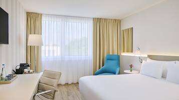 A hotel room with a bed, a desk, a chair, a small table with a vase of flowers, and a television.
