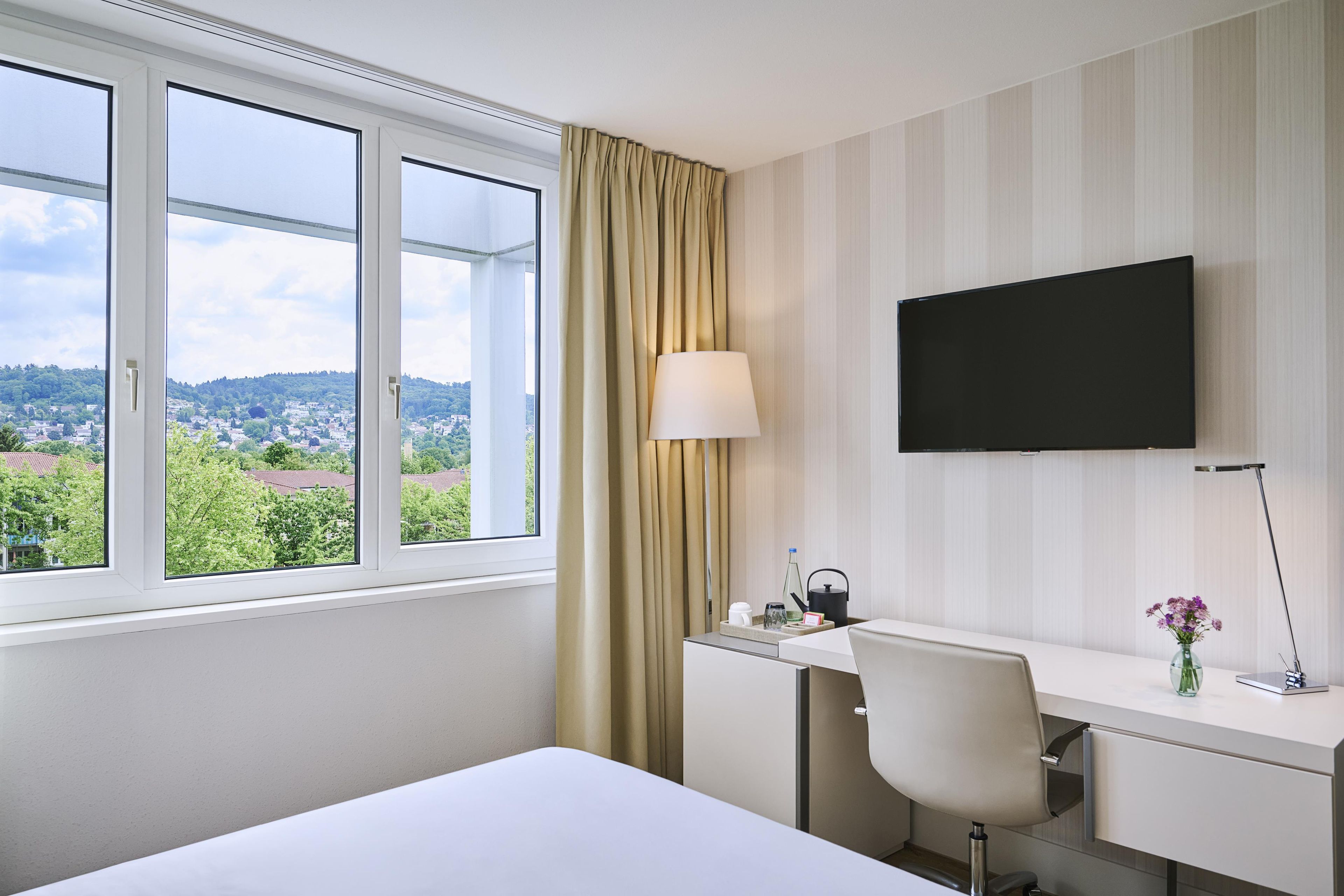 Ein Hotelzimmer mit einem Bett, einem Schreibtisch mit Stuhl, einem Fernseher, einem Fenster mit Vorhängen und Blick auf eine Stadtansicht.