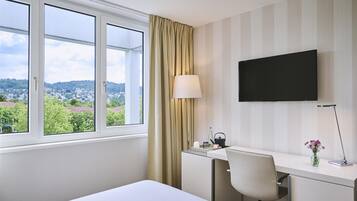 Ein Hotelzimmer mit einem Bett, einem Schreibtisch mit Stuhl, einem Fernseher, einem Fenster mit Vorhängen und Blick auf eine Stadtansicht.