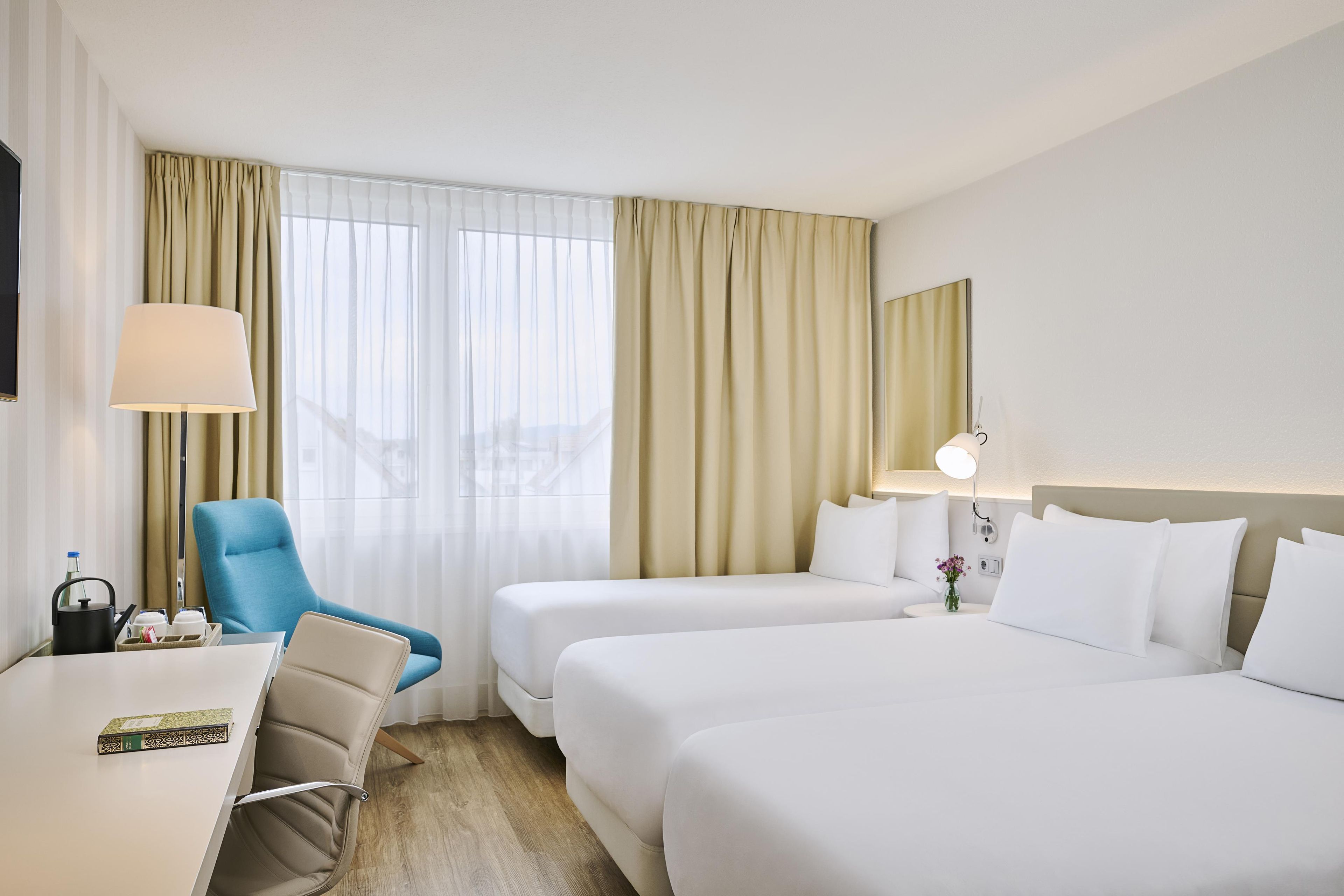 A hotel room with a desk, two beds, a chair, a lamp, and a view of the city through the window.
