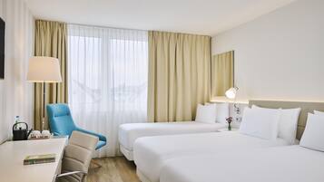 A hotel room with a desk, two beds, a chair, a lamp, and a view of the city through the window.