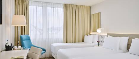 Habitación de hotel con escritorio, dos camas, una silla, lámpara y vista a la ciudad desde la ventana.