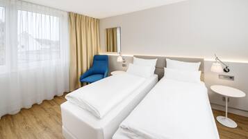 Habitación de hotel con dos camas individuales, una silla azul, una mesita redonda y un ventanal con cortinas de gasa.