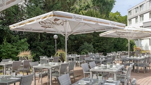 An outdoor cafe with wicker tables and chairs, covered by branded umbrellas.