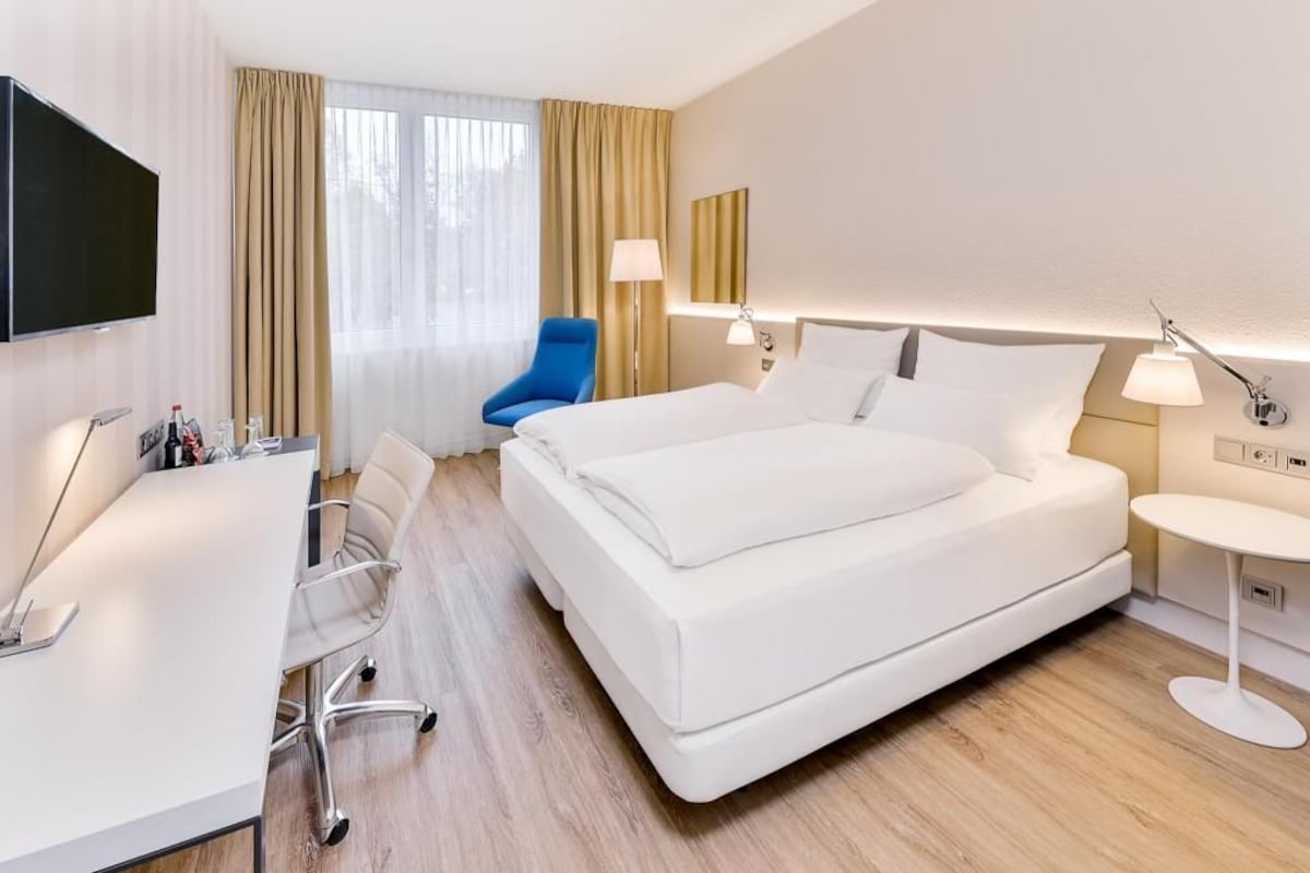 A hotel room with a bed, desk, chair, TV, and lamps.