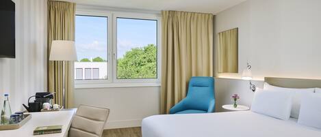 A hotel room with a bed, a desk, a chair, a window with curtains, and a flat-screen TV mounted on the wall.