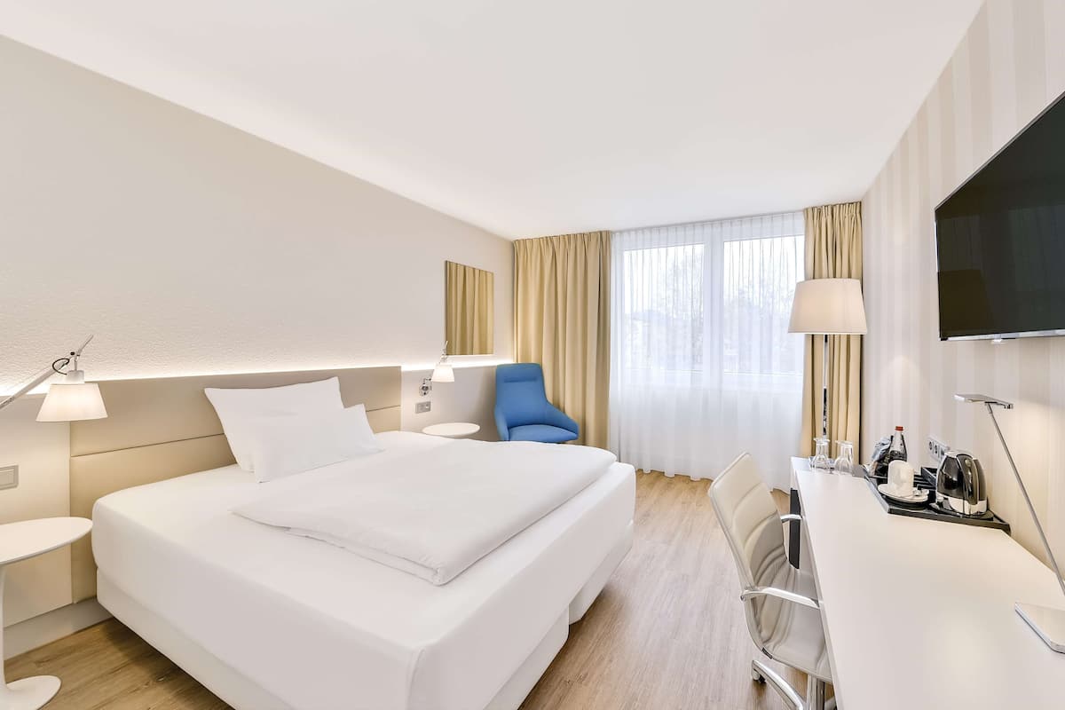 A hotel room with a large bed, a desk, a chair, a TV, and a window with curtains.