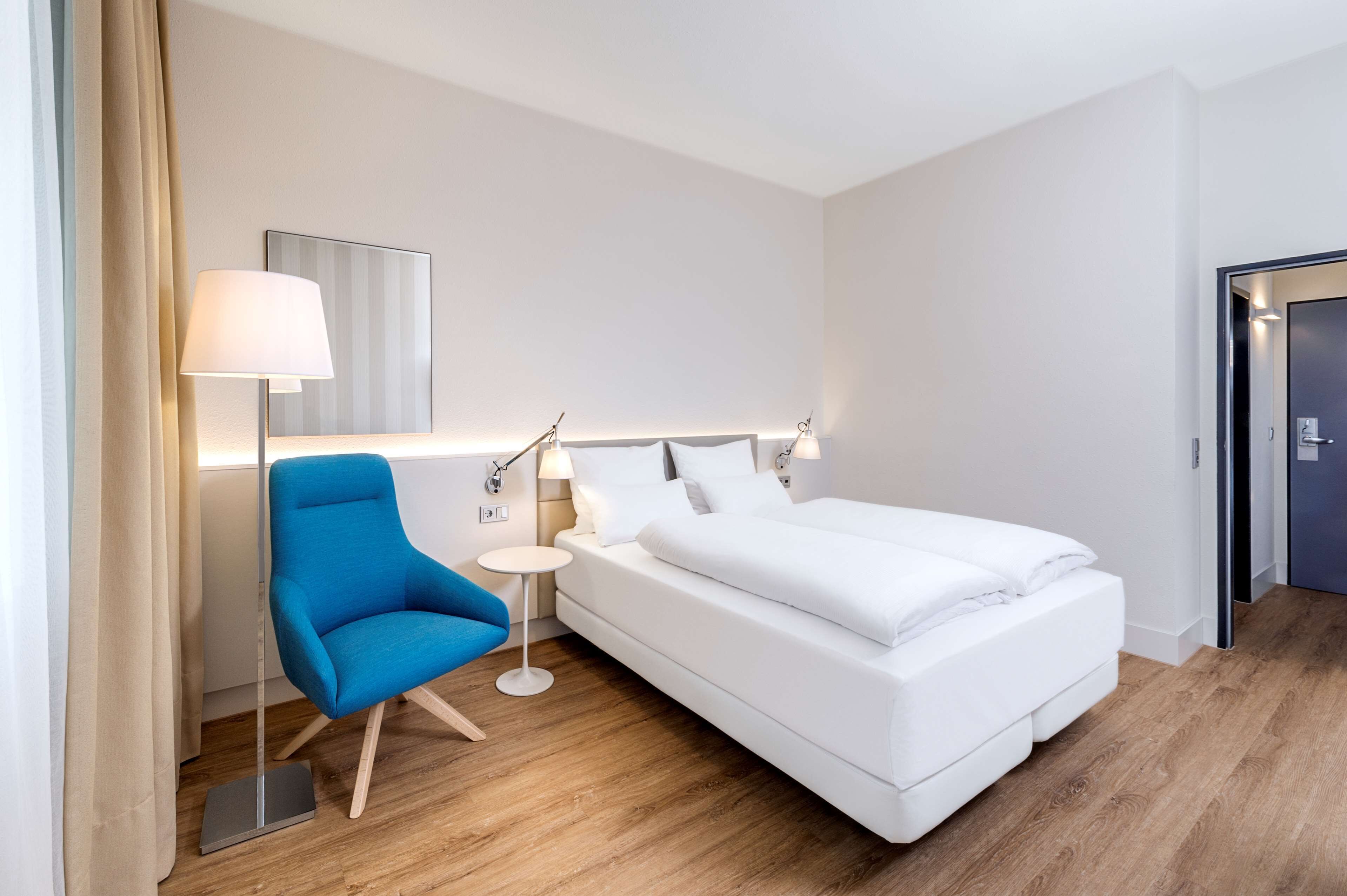 Een moderne hotelkamer met een groot bed, een blauwe fauteuil, een klein bijzettafeltje en een vloerlamp.