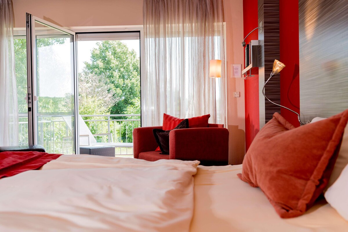 A hotel room with a bed, a red armchair, and a view of greenery outside.
