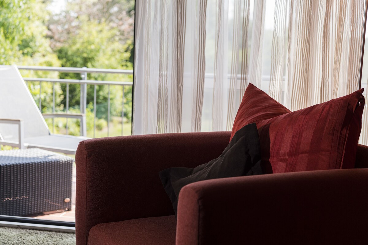 Un sofá rojo con un cojín negro en una habitación con un ventanal y vistas a zonas verdes.