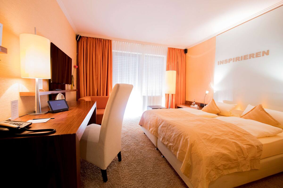 Ett hotellrum med en säng, ett skrivbord, en stol och ett stort fönster med gardiner.