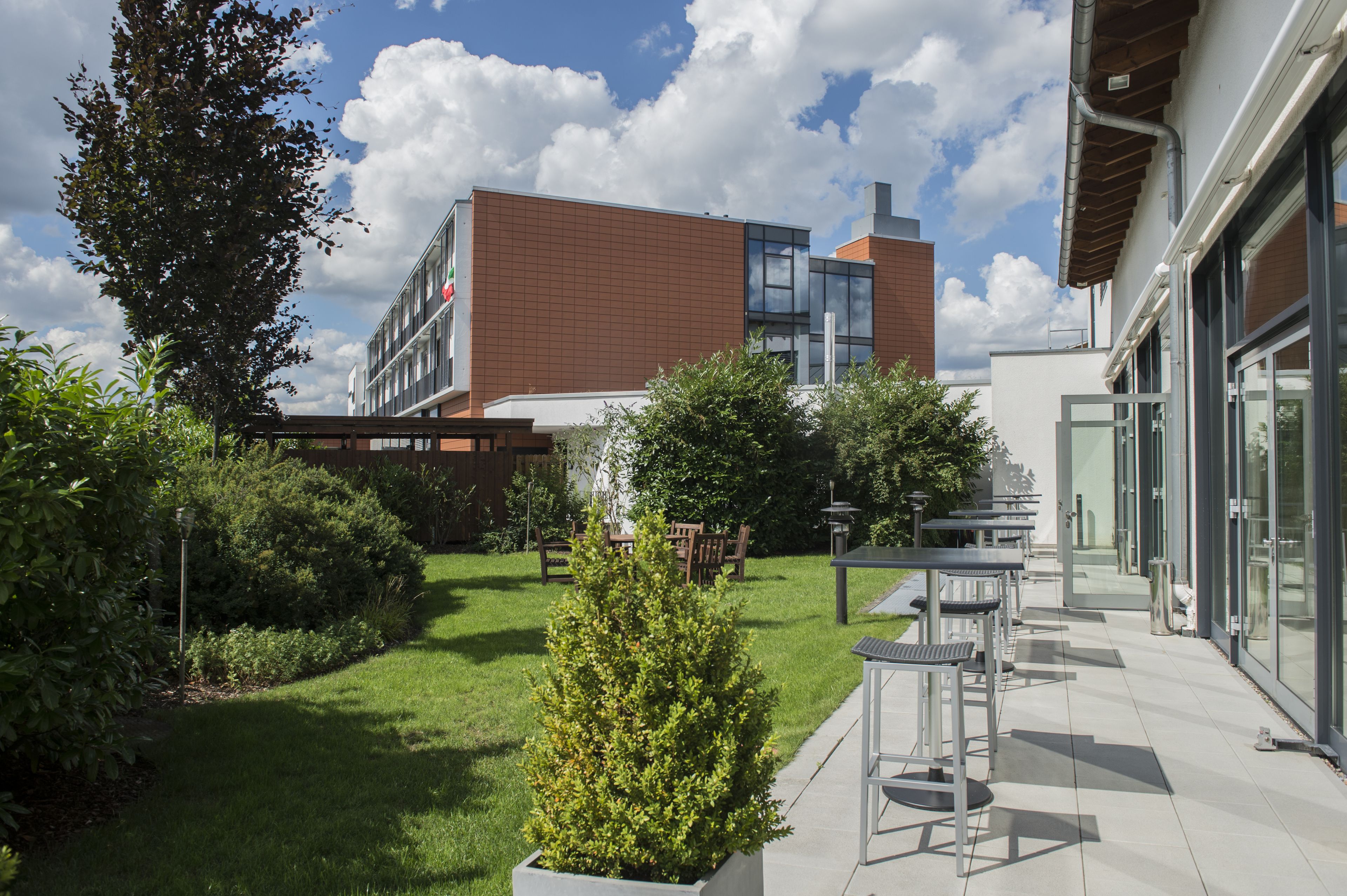 A modern building with a brick facade and large windows, surrounded by a well-maintained garden with grass, shrubs, and outdoor seating.