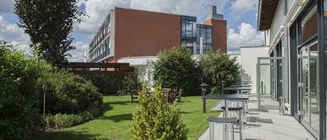 A modern building with a brick facade and large windows, surrounded by a well-maintained garden with grass, shrubs, and outdoor seating.
