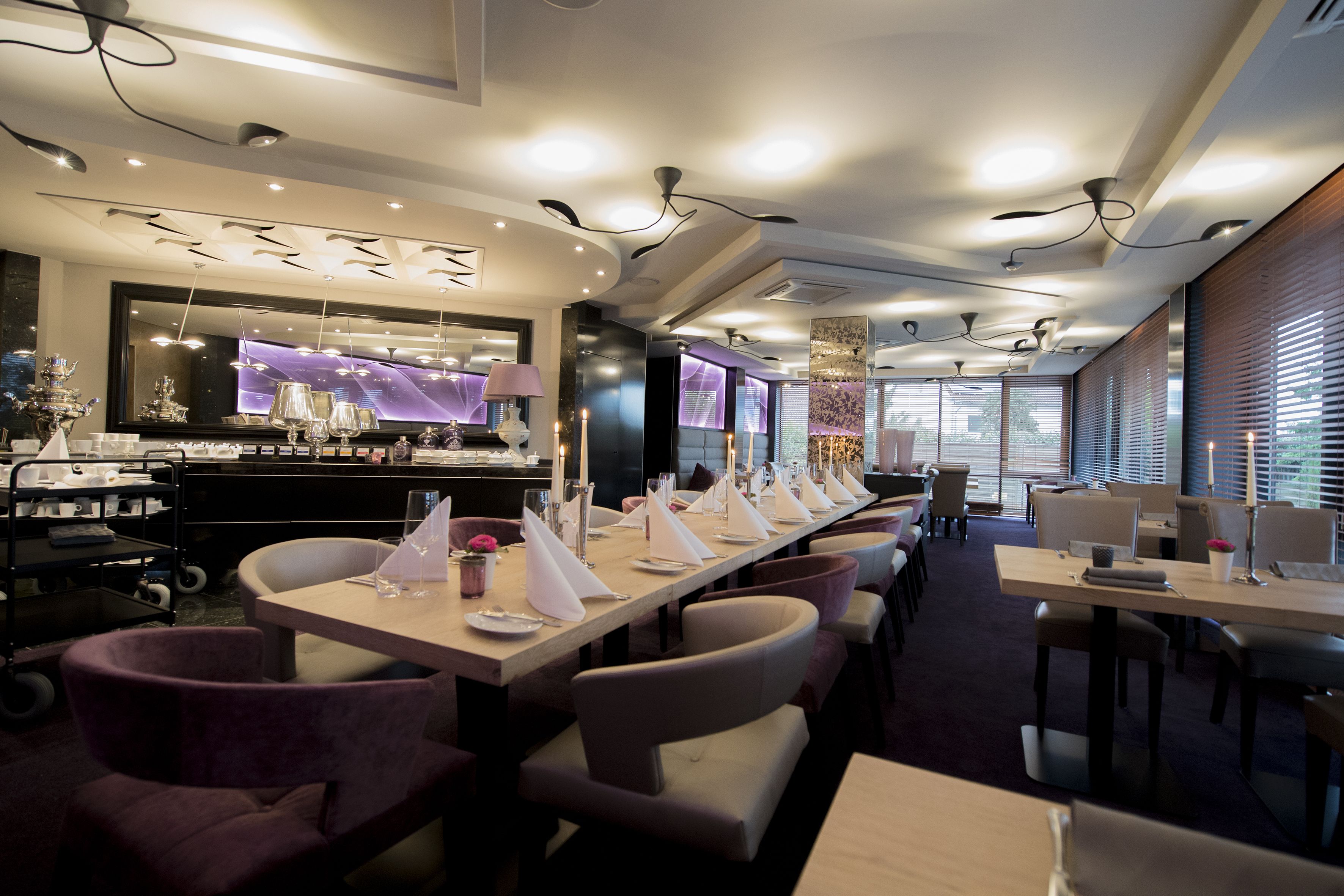 A modern restaurant with long tables, cushioned chairs, and a bar area with a large mirror and decorative items.