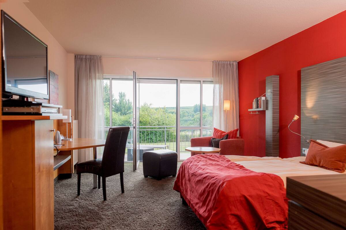 Ett hotellrum med en säng, ett skrivbord, en stol, ett litet bord och en balkong med utsikt.