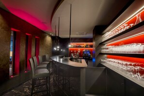 Zona de bar con mostrador curvo, taburetes y estante con varios vasos.