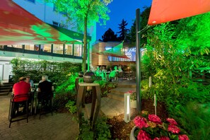An outdoor seating area with tables and chairs, surrounded by greenery and a building with large windows. - Atrium Hotel Mainz (Mainz)
