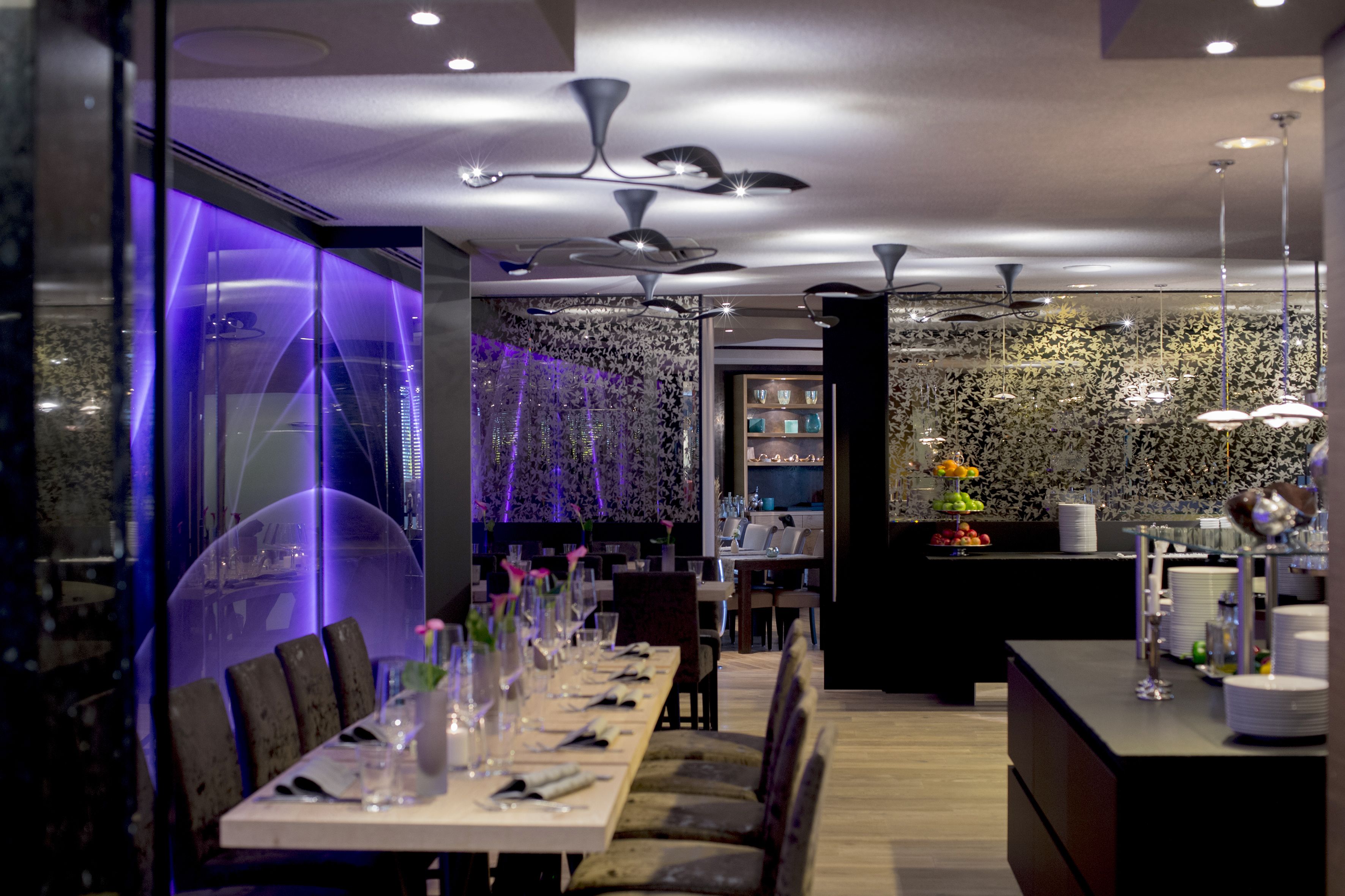 A modern restaurant with a long dining table set for a meal, a decorative wall with a floral pattern, and a bar area with a fruit display.